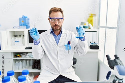 Middle age man working at scientist laboratory holding chemical products skeptic and nervous  frowning upset because of problem. negative person.