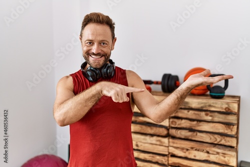 Middle age man wearing sportswear and headphones at the gym amazed and smiling to the camera while presenting with hand and pointing with finger. © Krakenimages.com