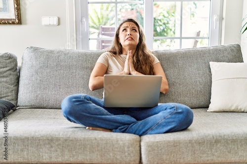 Beautiful young brunette woman sitting on the sofa using computer laptop at home begging and praying with hands together with hope expression on face very emotional and worried. begging.