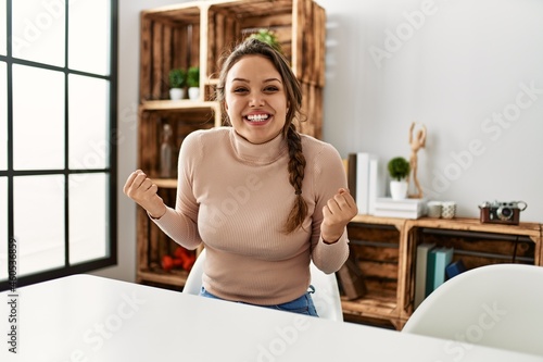 Young hispanic girl wearing casual clothes sitting on the table at home screaming proud  celebrating victory and success very excited with raised arms