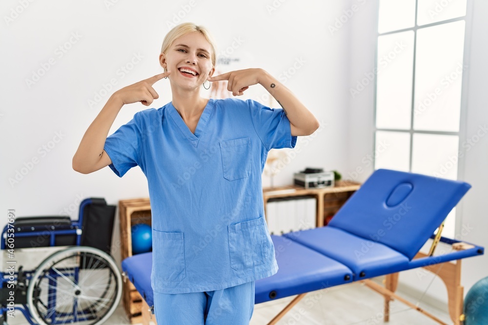 Beautiful caucasian physiotherapist woman working at pain recovery clinic smiling cheerful showing and pointing with fingers teeth and mouth. dental health concept. Beautiful caucasian physiotherapist woman working at pain recovery clinic smiling cheerful showing and pointing with fingers teeth and mouth. dental health concept.