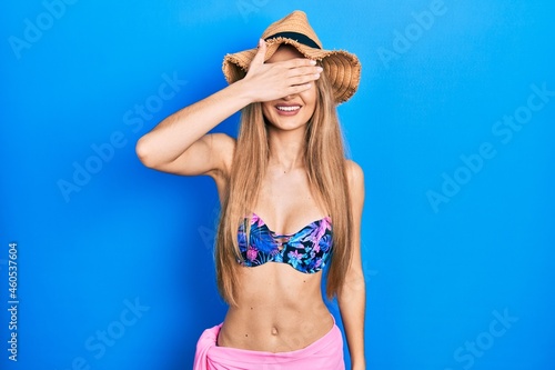 Young blonde girl wearing bikini and summer hat smiling and laughing with hand on face covering eyes for surprise. blind concept.