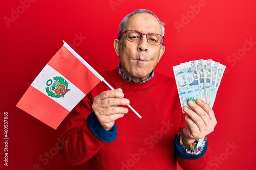 Handsome senior man with grey hair holding peru flag and peruvian sol banknotes making fish face with mouth and squinting eyes, crazy and comical.