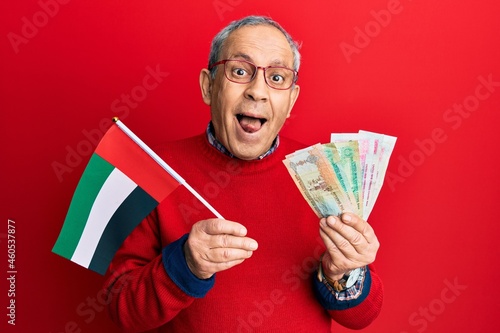 Handsome senior man with grey hair holding united arab emirates dirham banknotes sticking tongue out happy with funny expression.