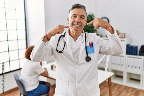 Middle age doctor man at the clinic with a patient smiling cheerful showing and pointing with fingers teeth and mouth. dental health concept.