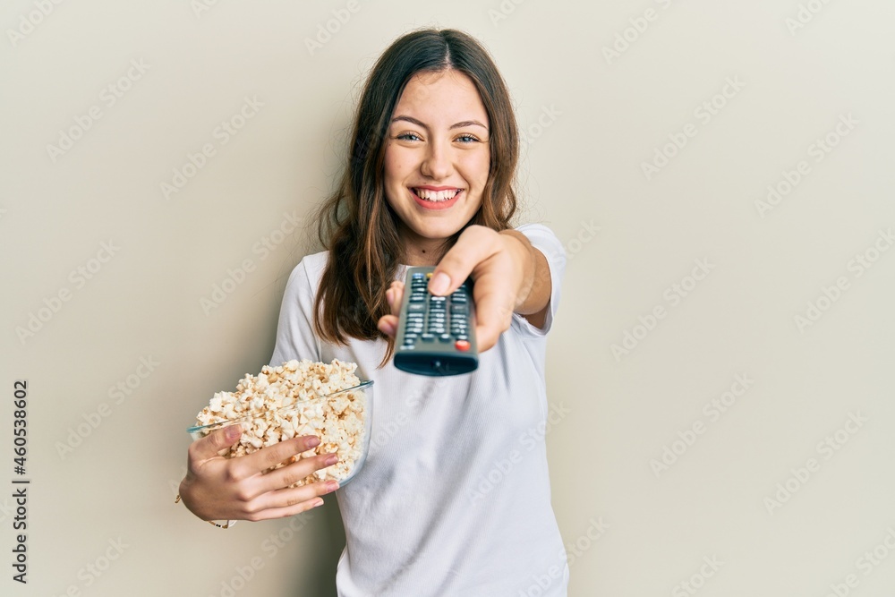 Young brunette woman eating popcorn using tv control smiling with a happy and cool smile on face. showing teeth. Young brunette woman eating popcorn using tv control smiling with a happy and cool smile on face. showing teeth.