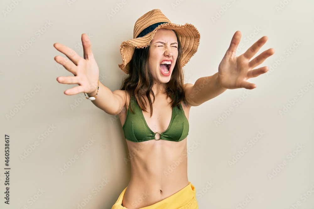 Young brunette woman wearing bikini and hat with open arms hugging angry and mad screaming frustrated and furious, shouting with anger looking up. Young brunette woman wearing bikini and hat with open arms hugging angry and mad screaming frustrated and furious, shouting with anger looking up.
