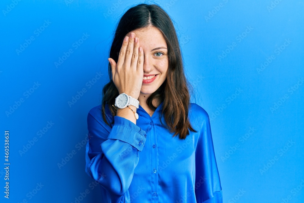 Young brunette woman wearing casual blue shirt covering one eye with hand, confident smile on face and surprise emotion. Young brunette woman wearing casual blue shirt covering one eye with hand, confident smile on face and surprise emotion.