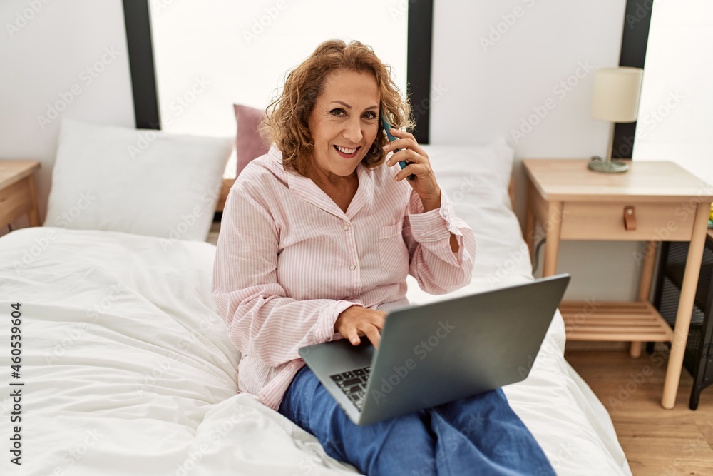 Middle age caucasian woman using laptop and talking on the smartphone sitting on the bed at bedroom.