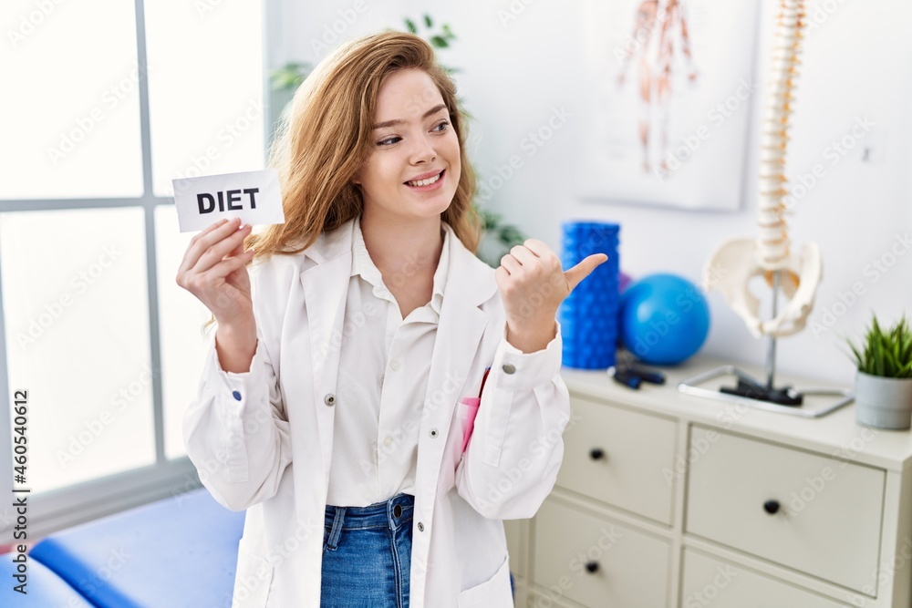 Young caucasian woman working at rehabilitation clinic holding diet banner pointing thumb up to the side smiling happy with open mouth Young caucasian woman working at rehabilitation clinic holding diet banner pointing thumb up to the side smiling happy with open mouth