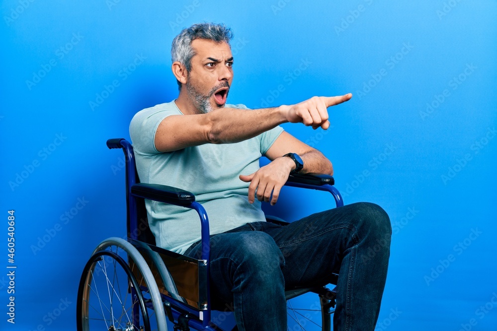 Fototapeta premium Handsome middle age man with grey hair sitting on wheelchair pointing with finger surprised ahead, open mouth amazed expression, something on the front