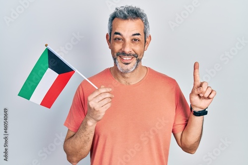 Handsome middle age man with grey hair holding kuwait flag smiling with an idea or question pointing finger with happy face, number one