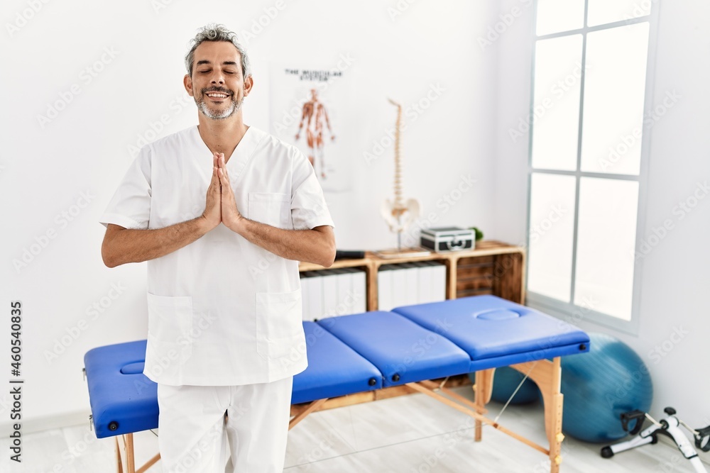 Middle age hispanic therapist man working at pain recovery clinic praying with hands together asking for forgiveness smiling confident. Middle age hispanic therapist man working at pain recovery clinic praying with hands together asking for forgiveness smiling confident.