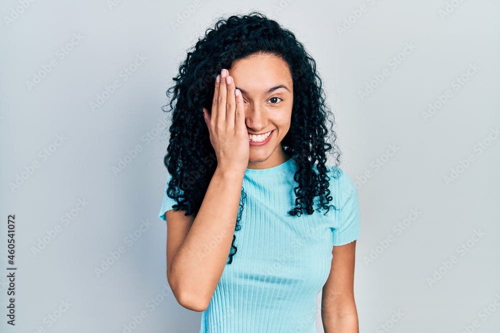 Young hispanic woman with curly hair wearing casual blue t shirt covering one eye with hand, confident smile on face and surprise emotion. Young hispanic woman with curly hair wearing casual blue t shirt covering one eye with hand, confident smile on face and surprise emotion.