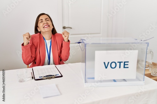 Beautiful middle age hispanic woman at political election sitting by ballot excited for success with arms raised and eyes closed celebrating victory smiling. winner concept.