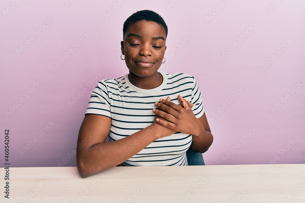Young african american woman wearing casual clothes sitting on the table smiling with hands on chest with closed eyes and grateful gesture on face. health concept.