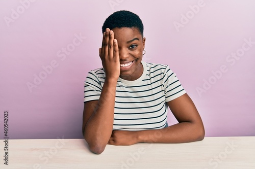 Young african american woman wearing casual clothes sitting on the table covering one eye with hand, confident smile on face and surprise emotion.