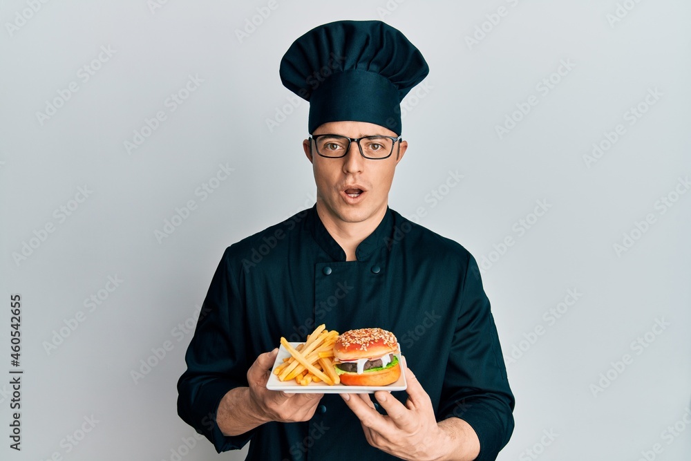 Handsome young man chef holding burger with fries afraid and shocked with surprise and amazed expression, fear and excited face. Handsome young man chef holding burger with fries afraid and shocked with surprise and amazed expression, fear and excited face.