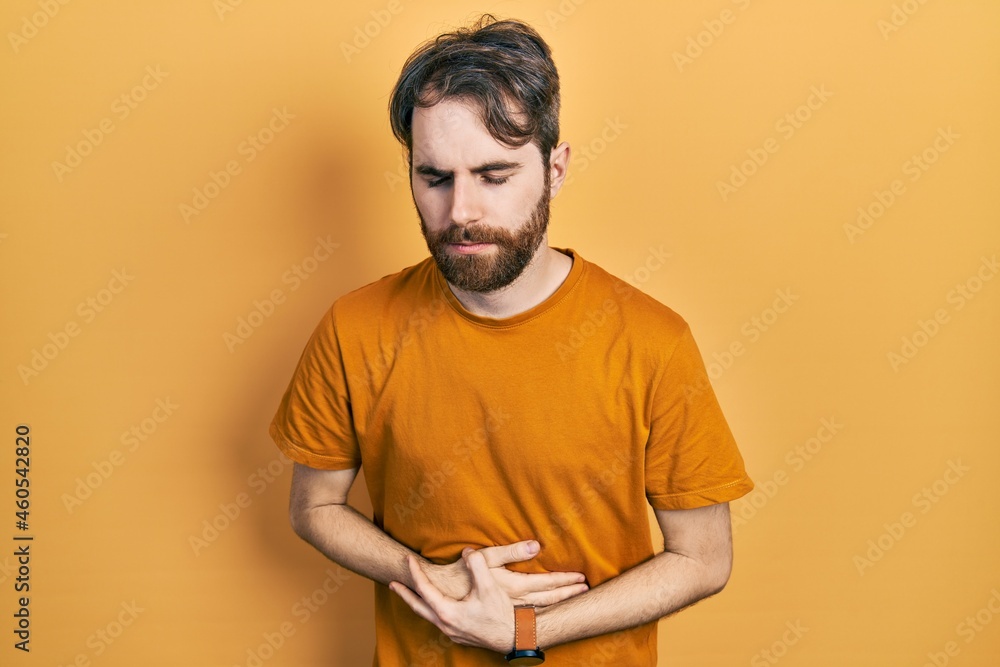 Caucasian man with beard wearing casual yellow t shirt with hand on stomach because indigestion, painful illness feeling unwell. ache concept. Caucasian man with beard wearing casual yellow t shirt with hand on stomach because indigestion, painful illness feeling unwell. ache concept.