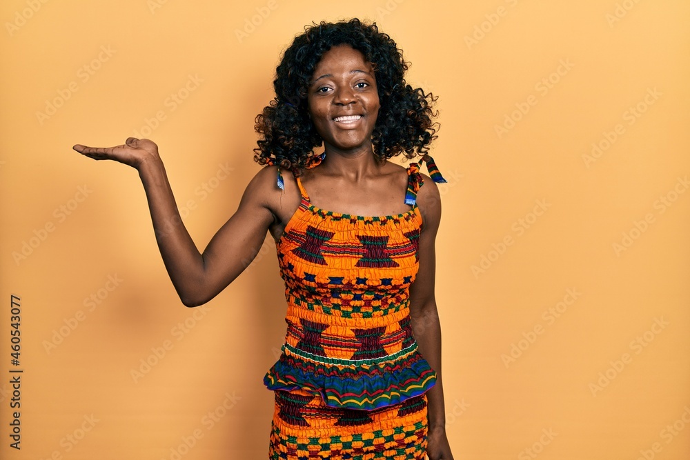 Young african american woman wearing traditional african clothes smiling cheerful presenting and pointing with palm of hand looking at the camera. Young african american woman wearing traditional african clothes smiling cheerful presenting and pointing with palm of hand looking at the camera.