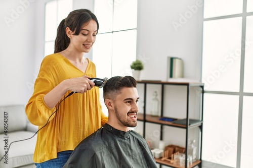 Young hispanic hairdresser woman cutting man s hair using electric razor machine at beauty center.