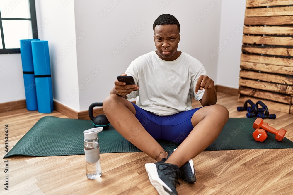 Young african man sitting on training mat at the gym using smartphone pointing down looking sad and upset, indicating direction with fingers, unhappy and depressed. Young african man sitting on training mat at the gym using smartphone pointing down looking sad and upset, indicating direction with fingers, unhappy and depressed.