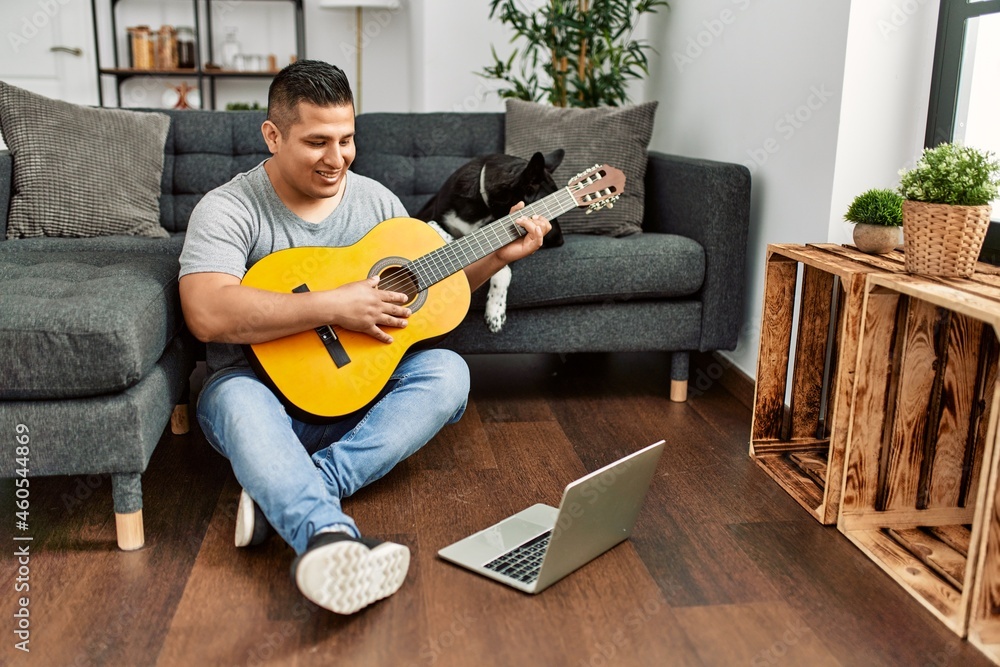 Young hispanic man having onlinr classical guitar lesson sitting on the floor with dog at home.