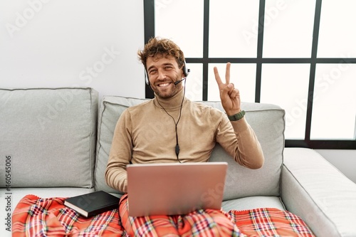 Young handsome man with beard wearing operator headset working from home smiling looking to the camera showing fingers doing victory sign. number two.