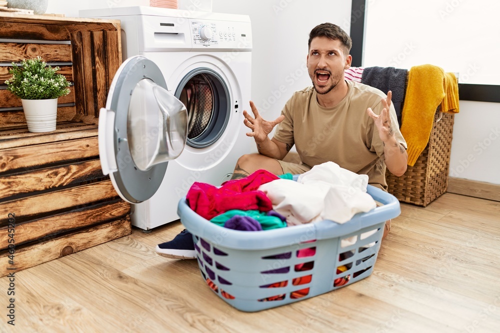 Young handsome man putting dirty laundry into washing machine crazy and mad shouting and yelling with aggressive expression and arms raised. frustration concept. Young handsome man putting dirty laundry into washing machine crazy and mad shouting and yelling with aggressive expression and arms raised. frustration concept.