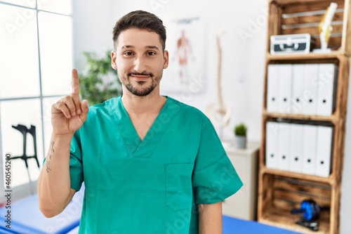 Young physiotherapist man working at pain recovery clinic showing and pointing up with finger number one while smiling confident and happy.
