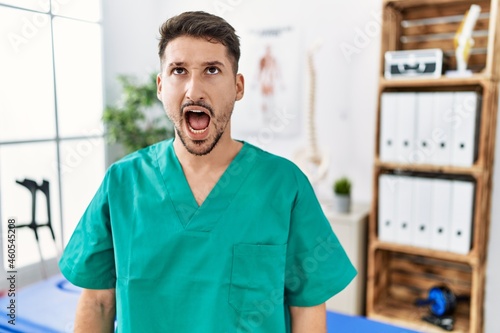 Young physiotherapist man working at pain recovery clinic angry and mad screaming frustrated and furious, shouting with anger. rage and aggressive concept.