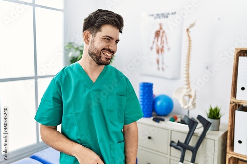 Young man with beard working at pain recovery clinic looking away to side with smile on face, natural expression. laughing confident.