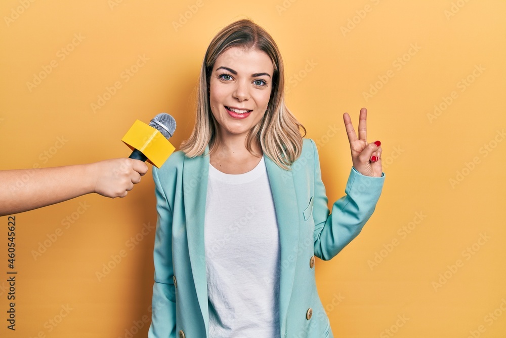 Beautiful caucasian woman being interviewed by reporter holding microphone smiling with happy face winking at the camera doing victory sign. number two. Beautiful caucasian woman being interviewed by reporter holding microphone smiling with happy face winking at the camera doing victory sign. number two.