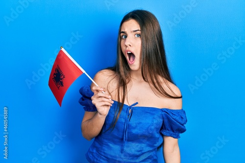 Young brunette teenager holding albania flag scared and amazed with open mouth for surprise  disbelief face