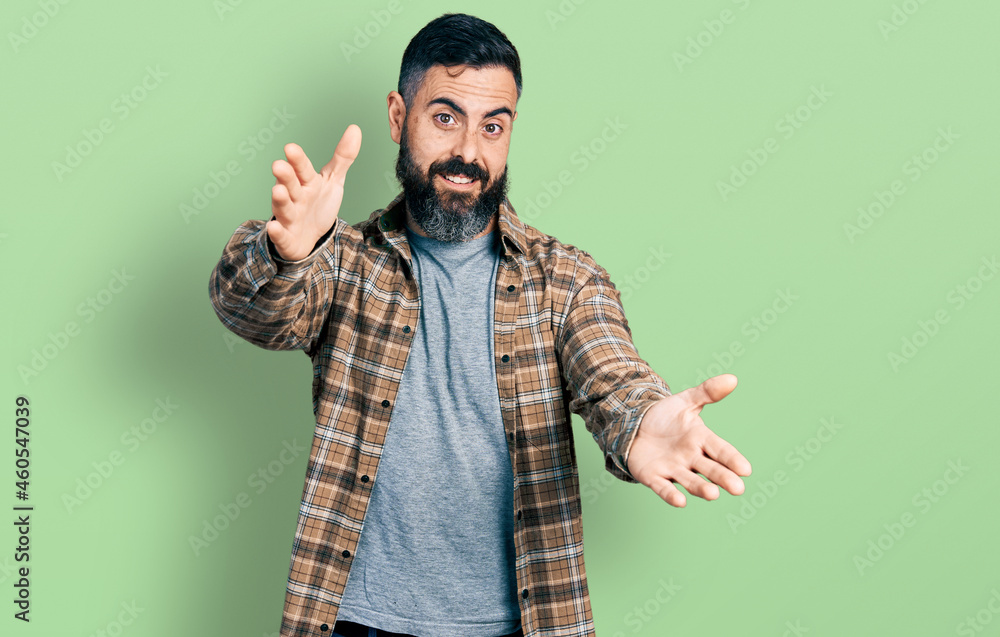 Hispanic man with beard wearing casual shirt looking at the camera smiling with open arms for hug. cheerful expression embracing happiness. Hispanic man with beard wearing casual shirt looking at the camera smiling with open arms for hug. cheerful expression embracing happiness.