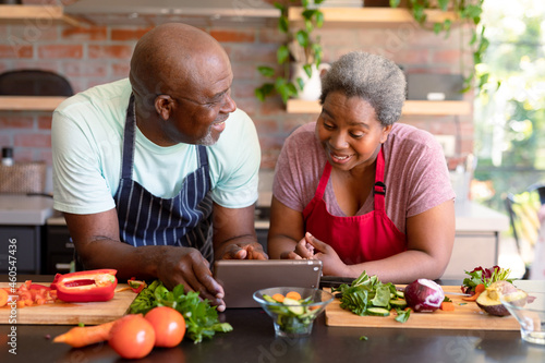 Happy african american senior couple cooking together in kitchen, using tablet