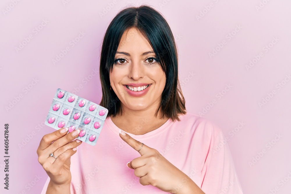 Young hispanic woman holding pills smiling happy pointing with hand and finger Young hispanic woman holding pills smiling happy pointing with hand and finger