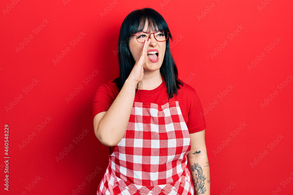 Young hispanic woman wearing cook apron and glasses shouting and screaming loud to side with hand on mouth. communication concept. Young hispanic woman wearing cook apron and glasses shouting and screaming loud to side with hand on mouth. communication concept.