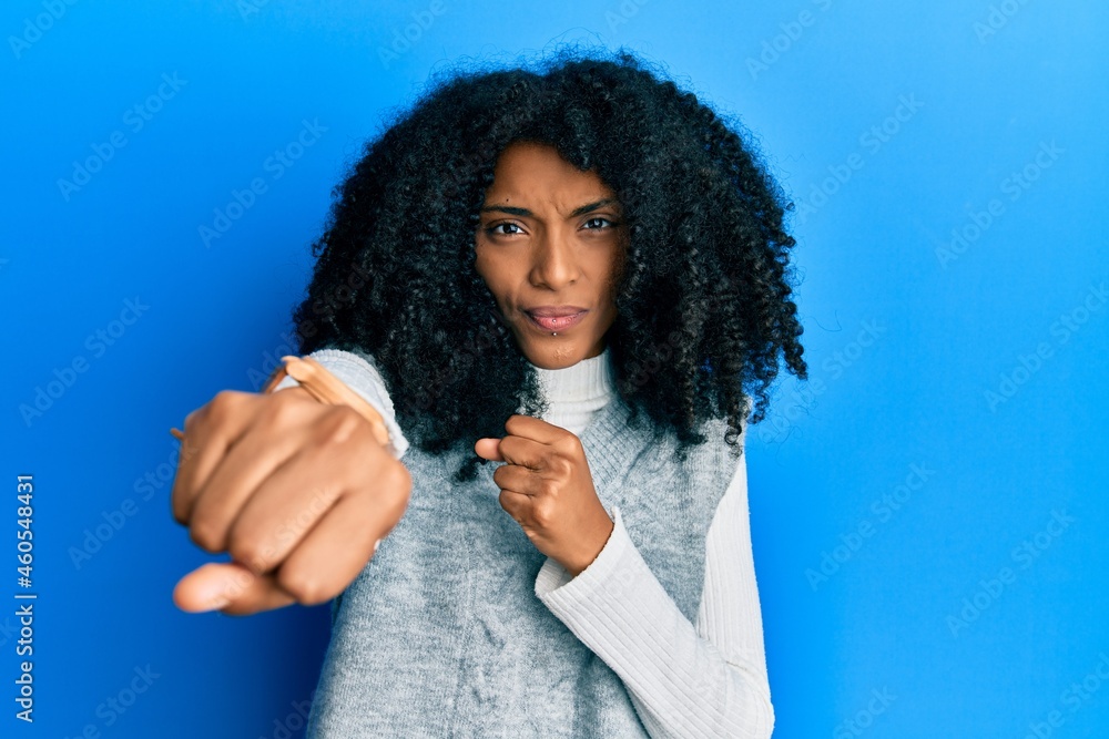 African american woman with afro hair wearing casual winter sweater punching fist to fight, aggressive and angry attack, threat and violence African american woman with afro hair wearing casual winter sweater punching fist to fight, aggressive and angry attack, threat and violence