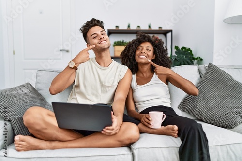 Young interracial couple using laptop at home sitting on the sofa smiling cheerful showing and pointing with fingers teeth and mouth. dental health concept.