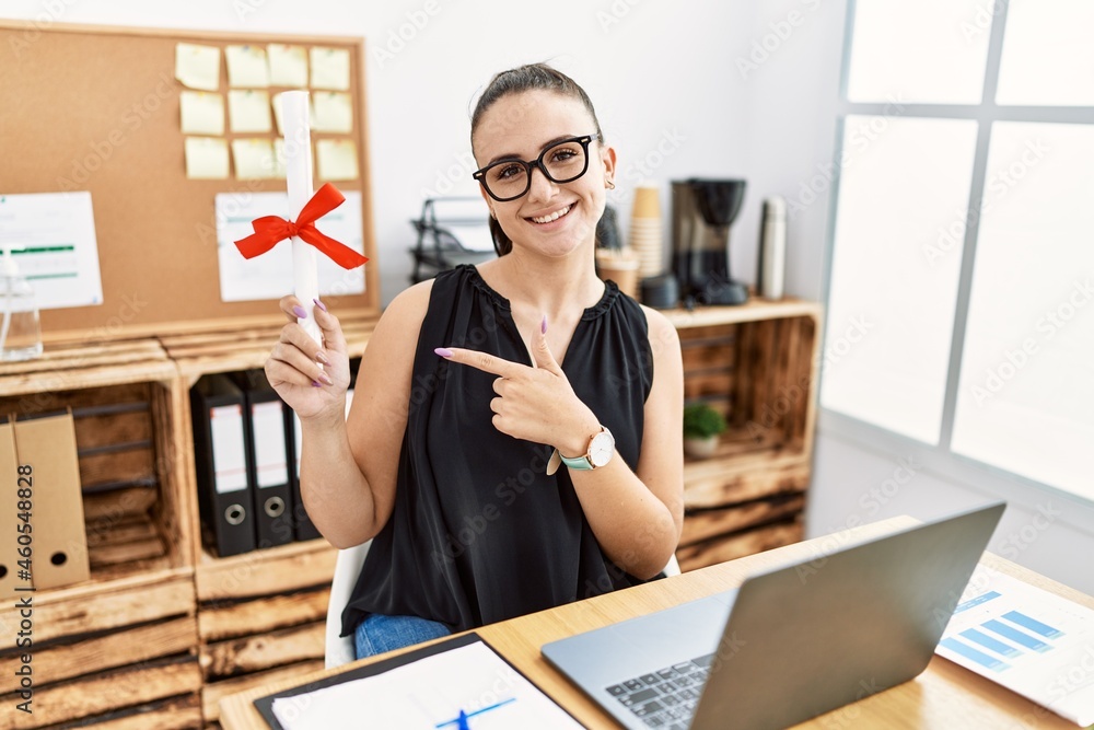 Young brunette woman holding graduate degree diploma at the office smiling happy pointing with hand and finger Young brunette woman holding graduate degree diploma at the office smiling happy pointing with hand and finger