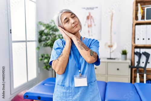 Middle age grey-haired woman wearing physiotherapist uniform at medical clinic sleeping tired dreaming and posing with hands together while smiling with closed eyes.