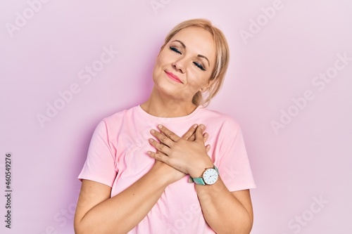 Young blonde woman wearing casual pink t shirt smiling with hands on chest with closed eyes and grateful gesture on face. health concept.