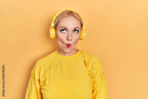 Young blonde woman listening to music using headphones making fish face with lips, crazy and comical gesture. funny expression.