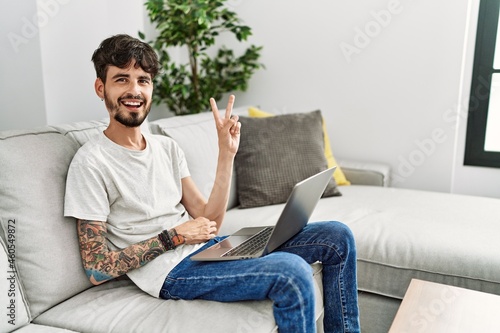 Hispanic man with beard sitting on the sofa smiling with happy face winking at the camera doing victory sign. number two.