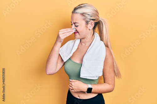 Young blonde girl wearing sportswear and towel smelling something stinky and disgusting, intolerable smell, holding breath with fingers on nose. bad smell