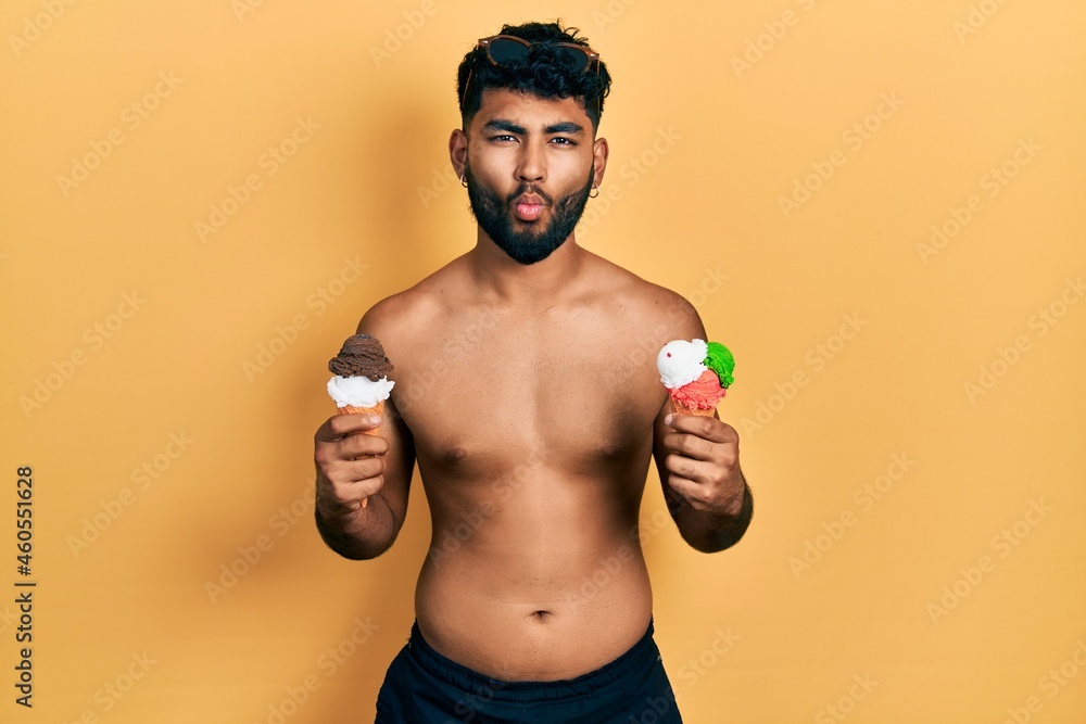 Arab man with beard wearing swimwear eating two ice cream cones making fish face with mouth and squinting eyes, crazy and comical. Arab man with beard wearing swimwear eating two ice cream cones making fish face with mouth and squinting eyes, crazy and comical.