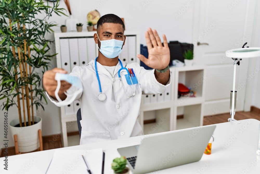 Young indian doctor offering safety mask with open hand doing stop sign with serious and confident expression, defense gesture Young indian doctor offering safety mask with open hand doing stop sign with serious and confident expression, defense gesture