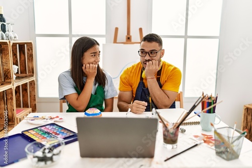 Young hispanic couple using laptop sitting on the table at art studio looking stressed and nervous with hands on mouth biting nails. anxiety problem.