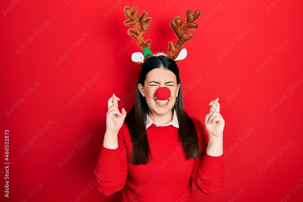 Young hispanic woman wearing deer christmas hat and red nose gesturing finger crossed smiling with hope and eyes closed. luck and superstitious concept.